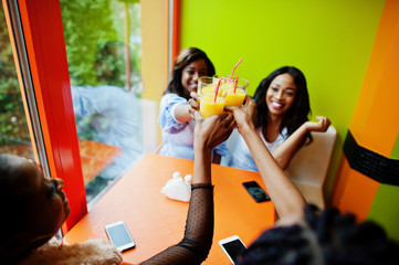 Four young african girls cheers with orange juices while sitting in bright colored fast food restaurant.
