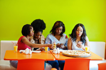 Four young african girls in bright colored fast food restaurant making photo of pizza in their mobile phones.