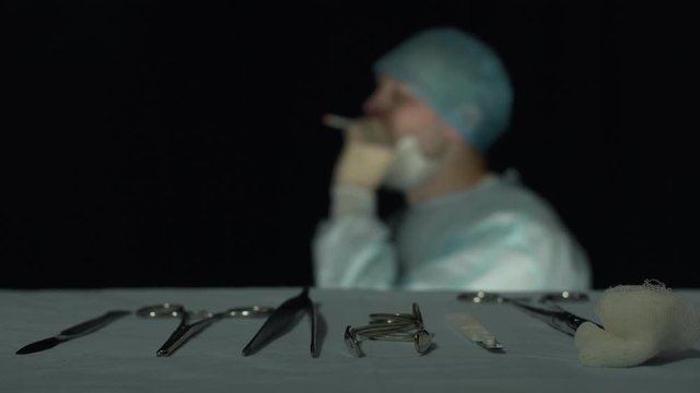 Tired surgeon smokes a cigarette after operation or procedure in hospital. Focus on surgical instruments such as scalpel, grasper, forcep, tweezer lie in the foreground