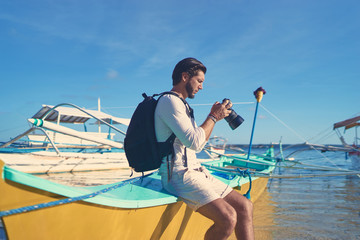 Photography and travel. Young man with rucksack taking photo with his camera enjoying beautiful tropical sea view on fishing beach. © Loginova