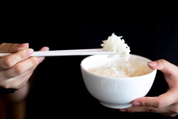 Hand of people using white chopsticks holding hot steamed rice in white bowl on dark background.