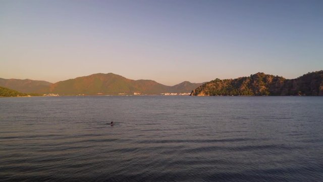 This shot depicts a panoramic view of a person swimming in Mediterranean sea during hot-summer surrounded by pine-clad mountains.