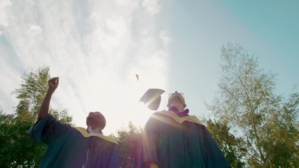 Two Male Happy Graduate Students in Gowns Throw Caps Up Outdoors after Graduation Ceremony or Diploma Party. 4K Slow Motion Medium Long Shot with Beautiful Sun Lens Flare