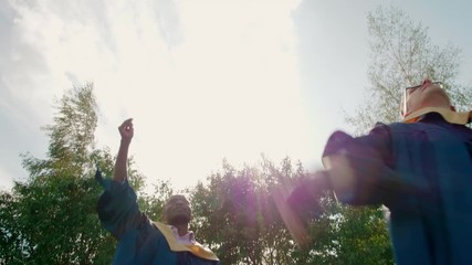 Two Male (African and Caucasian) Happy Graduate Students in Gowns Throw Caps Up Outdoors after Graduation Ceremony or Diploma Party. 4K Slow Motion Medium Long Shot with Beautiful Sun Lens Flare