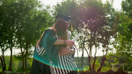 Excited Graduate Student in Gown and Cap with Diploma Hugs his Friend after Graduation Ceremony. 4K Slow Motion Static Medium Shot with Beautiful Sun Lens Flare