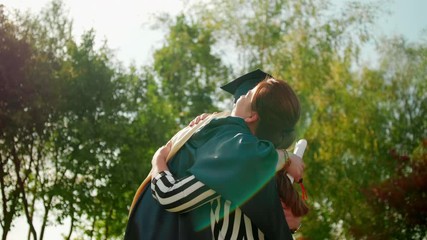 Excited Graduate Student in Gown and Cap with Diploma Hugs his Friend after Graduation Ceremony. 4K Slow Motion Medium Close-Up Shot with Beautiful Sun Lens Flare