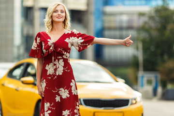 Photo of blonde in red dress stopping taxi on summer.