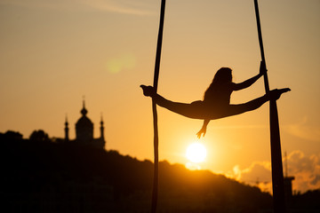 Silhouette of a flexible woman acrobat on aerial silk during a sunset on Kiev city background....