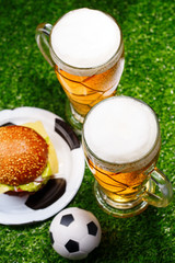 Two glasses of beer, hamburger and soccer ball on green grass.