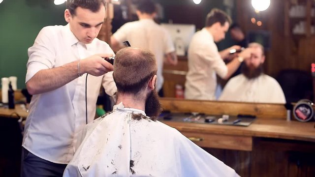 Young concentrated handsome barber working in hairdressing salon, doing confident moves with electric shaver. Man getting haircut, sitting back to camera and in front of mirror with blurred reflection