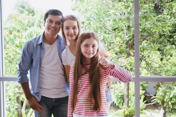 father and mother with daughter standing at window and raise hand at home, happy young family concept