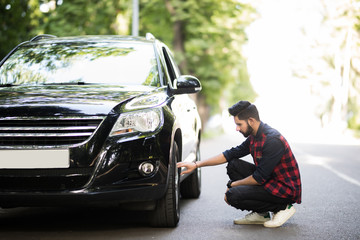 Cheerful young indian man has problems with the wheel of his car. He is kneeing and looking at it...