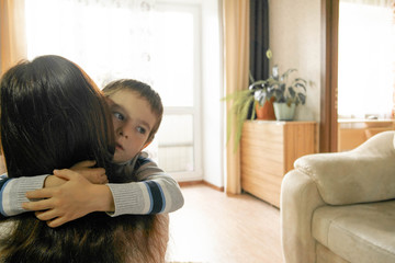 Mom hugs her son at home with light from the windows. Manifestation of family kindness and maternal love. Mutual understanding, sympathy and support of parents and children. Boy with sad emotions. 