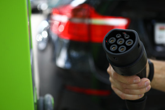 Focus on male hand holding modern charge device for cars. Automobile at filling station for electrocars. Environmental protection and newest transport technology concept. Blurred background