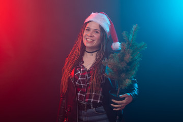Christmas, rock, studio, people concept - young woman in christmas hat with small christmas tree over the dark background