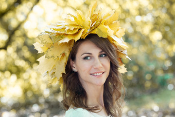 Closeup portrait of a beautiful smiling young brunette woman with a wreath of yellow maple leaves on her head. Golden autumn foliage of trees in the background