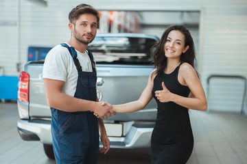 young handsome smiling mechanic and beautiful woman client shaking hands in gratitude for great service she shows like sign