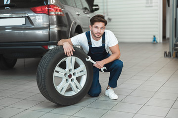 Obraz na płótnie Canvas young handsome mechanic wearing uniform working in car service department fixing flat tire looks pleased