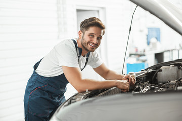 young handsome smiling mechanic wearing uniform fixing motor in car bonnet working in service department