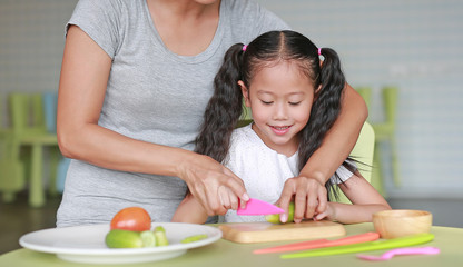 Close up Asian mom and daughter slicing vegetable on chopping board at play room. Mother teaches her little child girl to cooking.