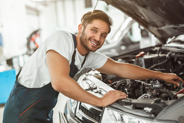 young smiling handsome mechanic in uniform fixing motor in car bonnet working in service center...
