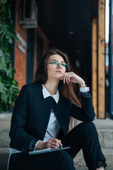 Young beautiful brunette girl, office clothing style, sitting on the street at a table in a cafe, street model posing style, makes notes in a notebook, talking on the phone