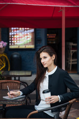 Young beautiful brunette girl, office clothing style, sitting on the street at a table in a cafe, street model posing style, makes notes in a notebook, talking on the phone