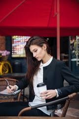 Young beautiful brunette girl, office clothing style, sitting on the street at a table in a cafe, street model posing style, makes notes in a notebook, talking on the phone
