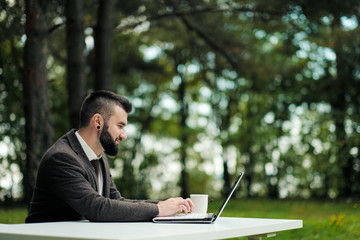 Young attractive business man in suit and tie sits at desk and works on computer outdoors. Drink coffee from white cup. Green trees, nature and park on background.