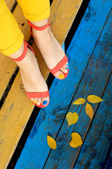 Multicolored autumn pedicure in orange sandals with yellow leaves on a blue wooden background close-up.