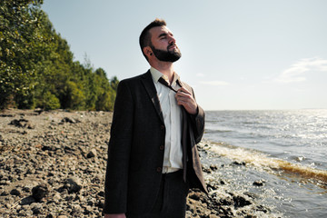 Young attractive male businessman in a brown suit with a briefcase stands on the seashore....