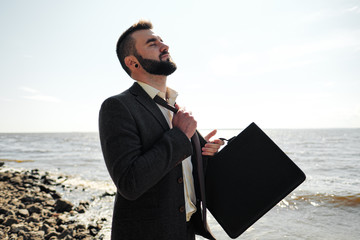 Young attractive male businessman in a brown suit with a briefcase stands on the seashore....