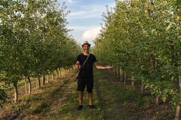 Smiling happy young man in hat wearing black t-shirt standing ampng the apple's garden and looking at the camera, showing ok sign with one hand, holding a prodact in other hand