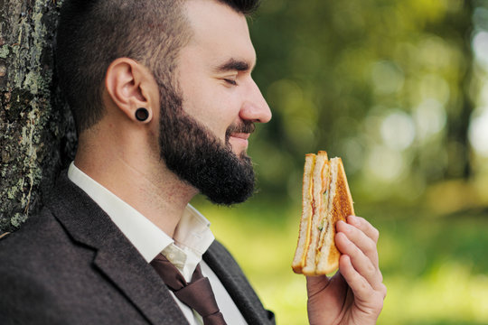 Young Attractive Business Man With Beard Sitting On Green Grass Under Tree And Resting In Park. Drinks Coffee And Eats A Sandwich. Relaxation, Tired Of Work, Lunch Break Outdoor.