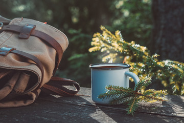 Enameled cup of coffee or tea, backpack of traveller on wooden board in summer forest outdoors.