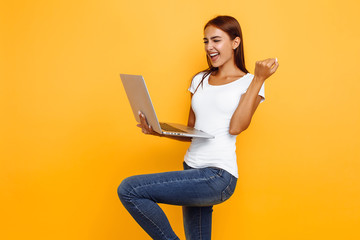 Portrait of excited young girl holding laptop and celebrating success isolated on yellow background