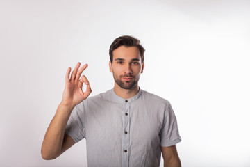 Fototapeta premium Young handsome bearded business man shows a finger okay and looking in camera while standing isolated in studio