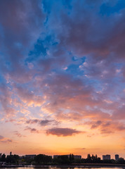 Cityscape of residential district under big sky at sunset. Buildings and vegetation at the front are almost silhouette and reflect on water