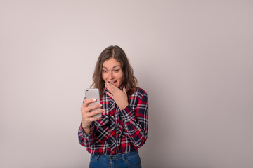 Surprised female in casual plaid shirt received good notifications on cellphone, standing isolated in studio