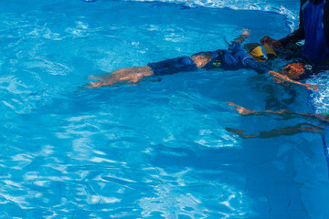Asian little boy age 5 years old learning basic to swim in the lesson of lying and floating technique on surface water with adult male swimmer and trainer in blue swimming pool background.