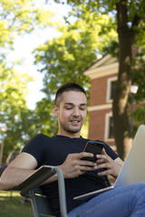 Fototapeta premium Fashionable hipster guy using messenger on cell telephone during distance work on laptop computer while sitting in park during summer weekend. Stylish man watching video on smartphone after webinar