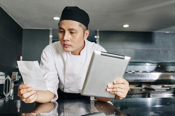 Asian young chef reading recipe of new dish and using digital tablet while standing in the kitchen