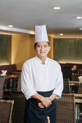 Portrait of Asian mature chef in uniform smiling at camera while standing at his restaurant