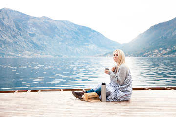 Woman on wooden pier by winter sea, mountains. Cozy picnic with coffee, hot beverages, tea or cocoa in thermos and mug, warm plaid, opened book. Girl is enjoying nature, wellbeing, reading on beach