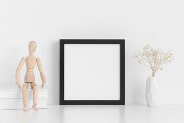 Black square frame mockup with books, manikin and a gypsophila in a vase on a white table.