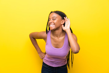 African American teenager girl with long braided hair over isolated yellow wall listening to...