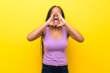 African American teenager girl with long braided hair over isolated yellow wall shouting and announcing something