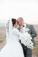 Beautiful newlyweds walking in the field admiring each other and kissing. Bridal couple, happy newlywed woman and man embracing. Loving wedding couple outdoor. Bridal bouquet of orchids 