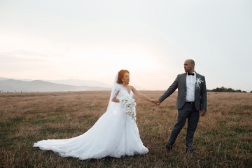 Romantic wedding couple having fun together outdoor at field. Groom and bride in a wedding dress going through the field on a background of blue sky at sunset. Wedding. Loving wedding couple
