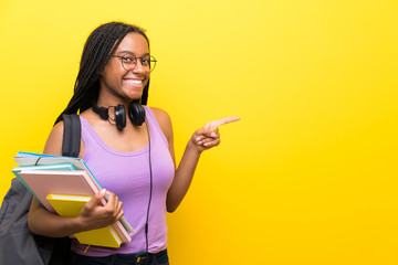 African American teenager student girl with long braided hair over isolated yellow wall pointing...
